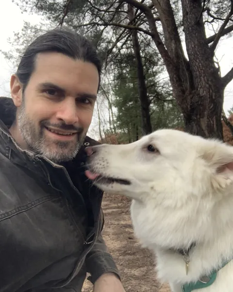 Rob Record smiling in a pine wood, with his white dog giving him a lick on the cheek